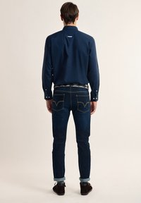 Camisa azul marino de botones, jeans de mezclilla oscura con bolsillos traseros visibles, mangas enrolladas y un cinturón con estampado. Zapatos marrones complementan el conjunto.