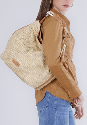 Donna che indossa una camicia marrone con bottoni e jeans, porta sulla spalla una grande borsa tote beige intrecciata con manici in pelle marrone chiaro.