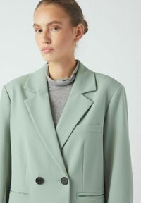 Mintgrønn dobbelspent blazer med svarte knapper, hakkede slag og en brystlomme, lagd over en grå høyhalset topp.