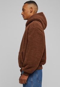 Rudas vilnos gobtuvas su tekstūruotu paviršiumi, reguliuojamu raišteliu ant gobtuvo ir įriebiomis rankovėmis. Matomas priekinis kengūros kišenė.