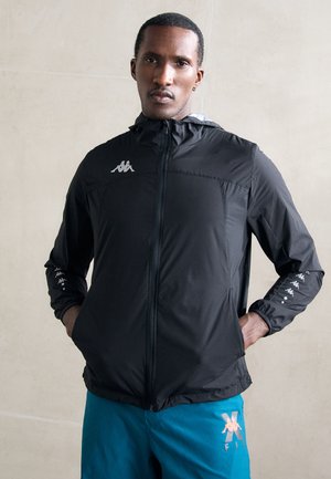 Veste zippée noire en matériau léger, dotée d'une capuche et de poignets élastiques. Logo en accent sur la poitrine et les manches. Associée à un short bleu.