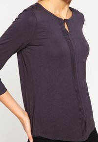 Ciemnofioletowa bluzka z długim rękawem i okrągłym dekoltem, z wycięciem z przodu oraz pionowymi fałdami wzdłuż środka. Miękka, lekka tkanina.