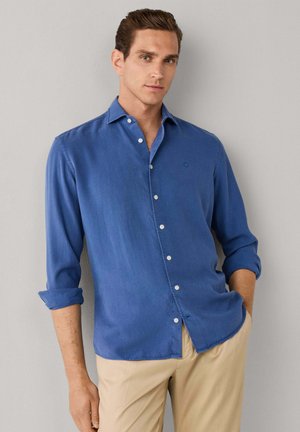 Hombre con camisa azul de manga larga abotonada con mangas remangadas, pantalones beige, de pie frente a una pared lisa gris claro.