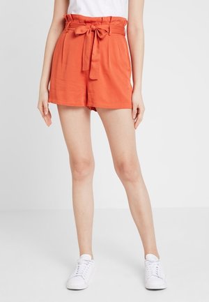 Frau trägt orangefarbene Paperbag-Shorts mit hoher Taille und gebundenem Gürtel, weißes Hemd und weiße Turnschuhe, steht vor einem schlichten weißen Hintergrund.