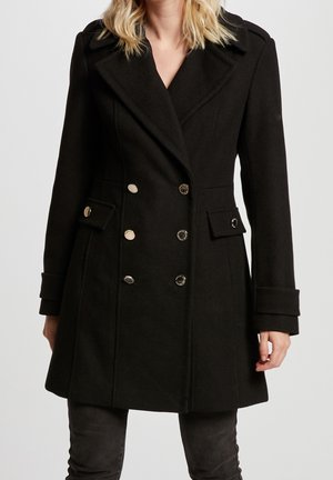 Vrouw draagt een zwart double-breasted wollen jas met zilverkleurige knopen en klepzakken, gecombineerd met donkere broek tegen een effen achtergrond.