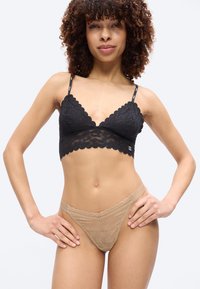 Femme portant un bralette en dentelle noire et une culotte beige en dentelle, debout les mains sur les hanches devant un fond uni.