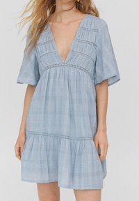 Světlomodré šaty s krátkým rukávem, hlubokým výstřihem do V, s nabíranou texturou a vrstveným lemem. Obsahují dekorativní krajkové akcenty.