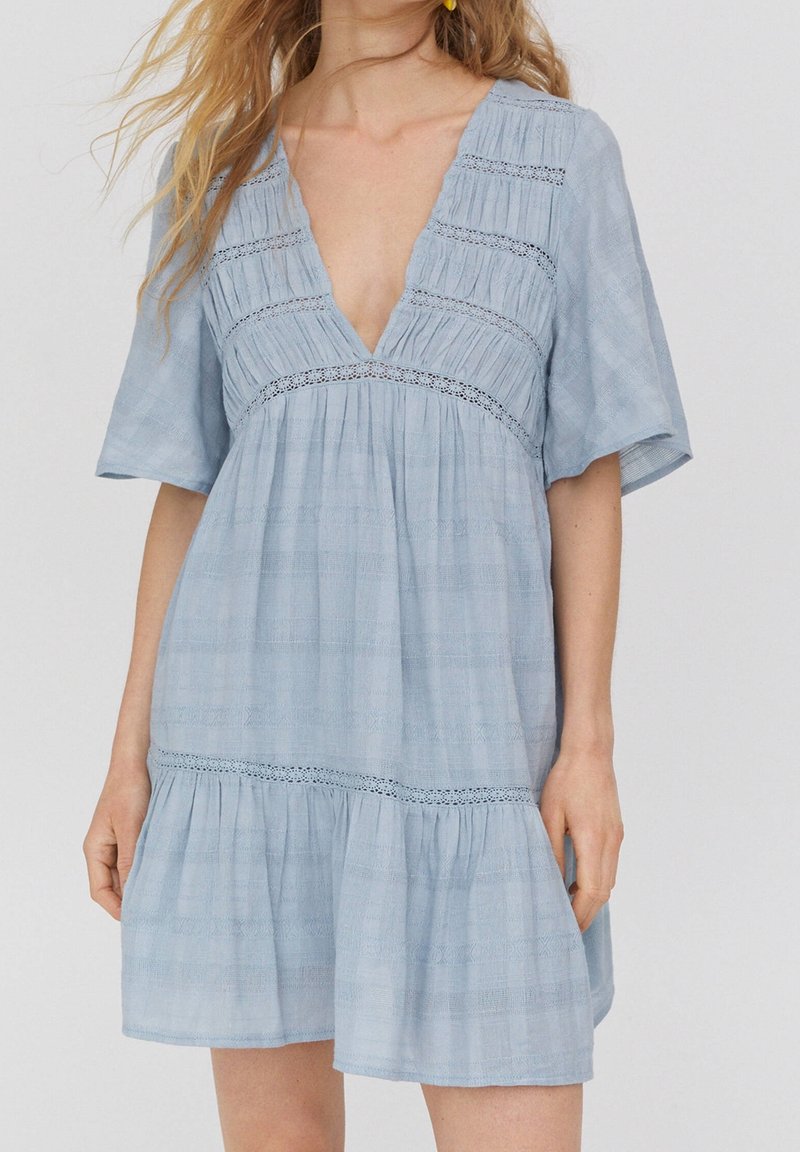 Světlomodré šaty s krátkým rukávem, hlubokým výstřihem do V, s nabíranou texturou a vrstveným lemem. Obsahují dekorativní krajkové akcenty.