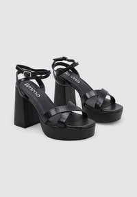 Sandalias de tacón alto negras con correas texturizadas, suela de plataforma y tacón cuadrado. Cuentan con correas ajustables en el tobillo y un acabado suave.