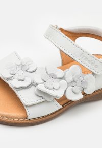 Valkoiset nahkaiset lasten sandaalit, joissa on kolme kerroksista kukka koristeita hihnassa ja ruskea pohja, jossa on ompeluyksityiskohtia.
