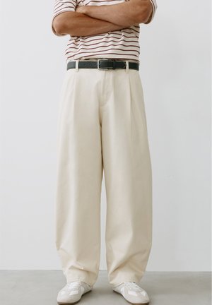 Personne debout, les bras croisés, portant un pantalon beige à jambes larges, une ceinture noire, des baskets blanches et une chemise rayée à manches courtes.