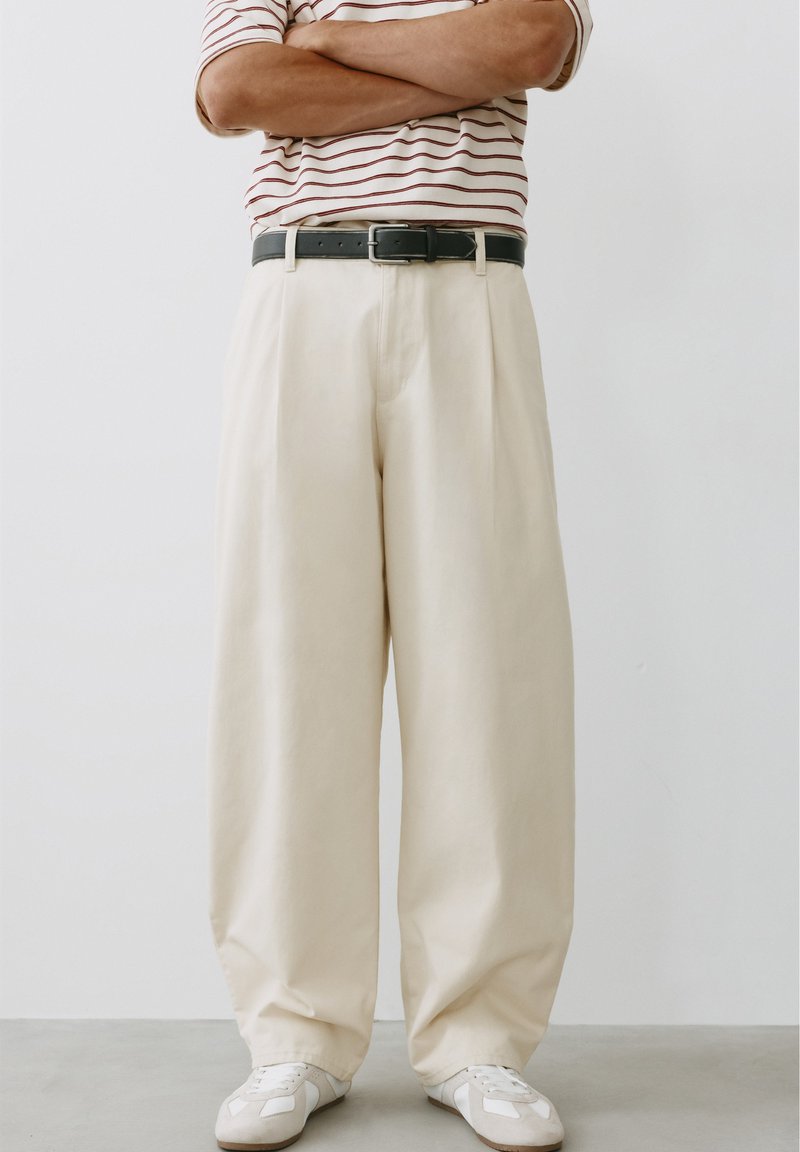 Personne debout, les bras croisés, portant un pantalon beige à jambes larges, une ceinture noire, des baskets blanches et une chemise rayée à manches courtes.
