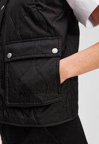 Crni kviltani prsluk s dva prednja džepa sa srebrnim dugmadi na zatvaranju i izvezenim detaljem "KARL LAGERFELD JEANS" na rubu.