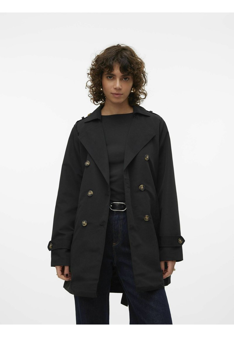 Black double-breasted trench coat with large buttons, wide lapels, and slightly oversized fit, paired with a black top and dark jeans.