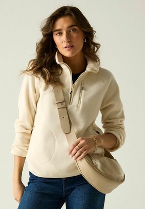 Donna con pile beige con zip a quarto, jeans blu e borsa beige a tracolla, in piedi davanti a uno sfondo chiaro e neutro.