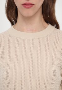 Gros plan d'une personne portant un pull en maille beige texturé, montrant les lèvres, le cou et une cicatrice légère à l'avant du cou.