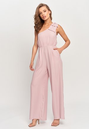 Kobieta o falowanych włosach, ubrana w różowy, różany, bezrękawnikowy kombinezon z szerokimi nogawkami i kokardą na jednym ramieniu, pozująca z jedną ręką w kieszeni.