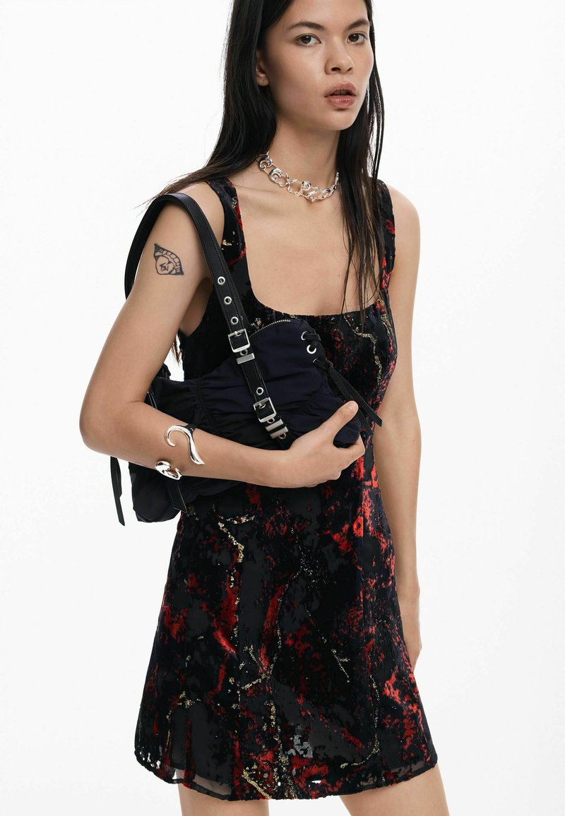 Must kleit punaste, kuldsete ja hõbedaste mustritega, millel on ruudukujuline kaelus. Model hoiab tumesinist kotti, millel on rihmad ja hõbedased detailid.