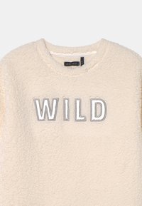 Sudadera de forro polar color crema con cuello redondo y la palabra "WILD" bordada en hilo metálico plateado en el pecho.