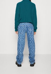 Niebieskie dżinsy z wzorem łączącym motywy kratki i abstrakcyjne, o luźnym kroju i widocznych detalach szwów. Noszone z zielonym swetrem.