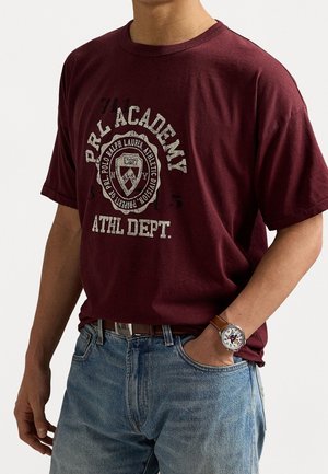 Personne portant un t-shirt graphique PRL Academy bordeaux, un jean bleu, une ceinture marron et une montre-bracelet avec un bracelet en cuir marron.