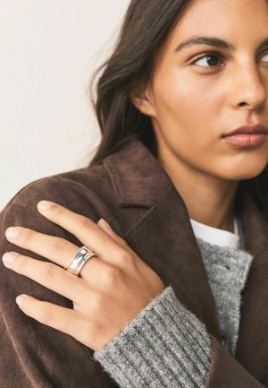 Woman with light brown skin wearing a brown coat and gray sweater, showing a silver wide band ring on her left hand resting on her shoulder.