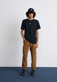 Crna Nike majica s bijelim logom, smeđi flis hlače s bočnim šavovima i crne tenisice s plavim naglascima. Model pozira na jednoličnoj pozadini.