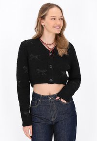 Cardigan corto nero in morbido tessuto a maglia, con scollo a V, chiusura a bottoni e motivi a fiocco testurizzati su tutto il capo. Abbinato a jeans blu.