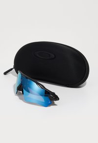 Czarne sportowe okulary przeciwsłoneczne z niebieskimi soczewkami, aerodynamiczny design, teksturowane uchwyty, obok czarnego pokrowca ochronnego z zamkiem.