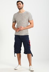 Stripete kortermet beige og marineblå T-skjorte, parret med marineblå cargo shorts med lommer og røde detaljer, brukt med hvite joggesko.
