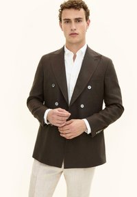 Mężczyzna w ciemnobrązowej dwurzędowej marynarce, białej koszuli zapinanej na guziki i jasnobeżowych spodniach, stojący na tle jednolitego tła.