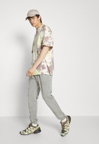 Tricou din bumbac tie-dye în nuanțe de verde, galben și maro, asortat cu pantaloni cargo gri deschis și adidași cu accente verzi și galbene.