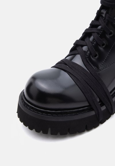 Μαύρο γυαλιστερό μποτάκι μάχης με παχύ πέλμα λάγκ τρακ, μαύρα κορδόνια που διασταυρώνονται και τυλίγονται γύρω από την περιοχή των δαχτύλων.