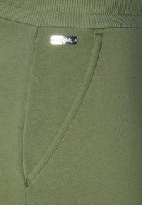 Olijfgroene joggingbroek met geribde tailleband en zijzak, voorzien van een klein zilveren logo-label. De stof is soepel en rekbaar.