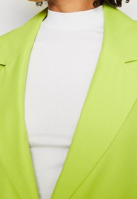 Jasnozielona marynarka o strukturze z wyraźnie zaznaczonymi ramionami, noszona na białym topie z wysokim kołnierzem. Gładka faktura materiału i wyraźnie zarysowany kołnierz.