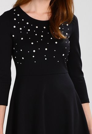 Zwarte jurk met driekwart mouwen, versierd met decoratieve witte parels in verschillende groottes op het lijfje. Gladde stof, aansluitende taille, uitlopende rok.