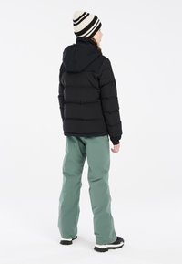 Veste d'hiver noire avec capuche, associée à un pantalon vert clair. Le mannequin porte un bonnet rayé et des bottes de ski noires, affichant une tenue décontractée.