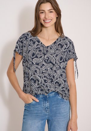 Kvinde smiler iført en navyblå paisley kortærmet bluse med sidebindede ærmer og blå jeans, med den ene hånd i lommen.