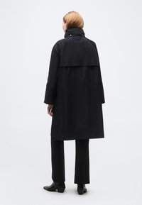 Άτομο με κοντά ξανθά μαλλιά που φοράει μακρύ μαύρο παλτό, μαύρο παντελόνι και μαύρες ψηλοτάκουνες μπότες, στέκεται με την πλάτη στο φόντο.