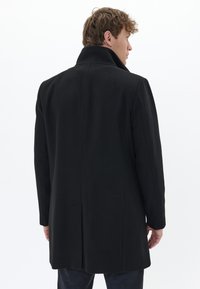 Homme avec des cheveux bouclés courts portant un manteau noir avec un col relevé, de dos contre un fond blanc uni.