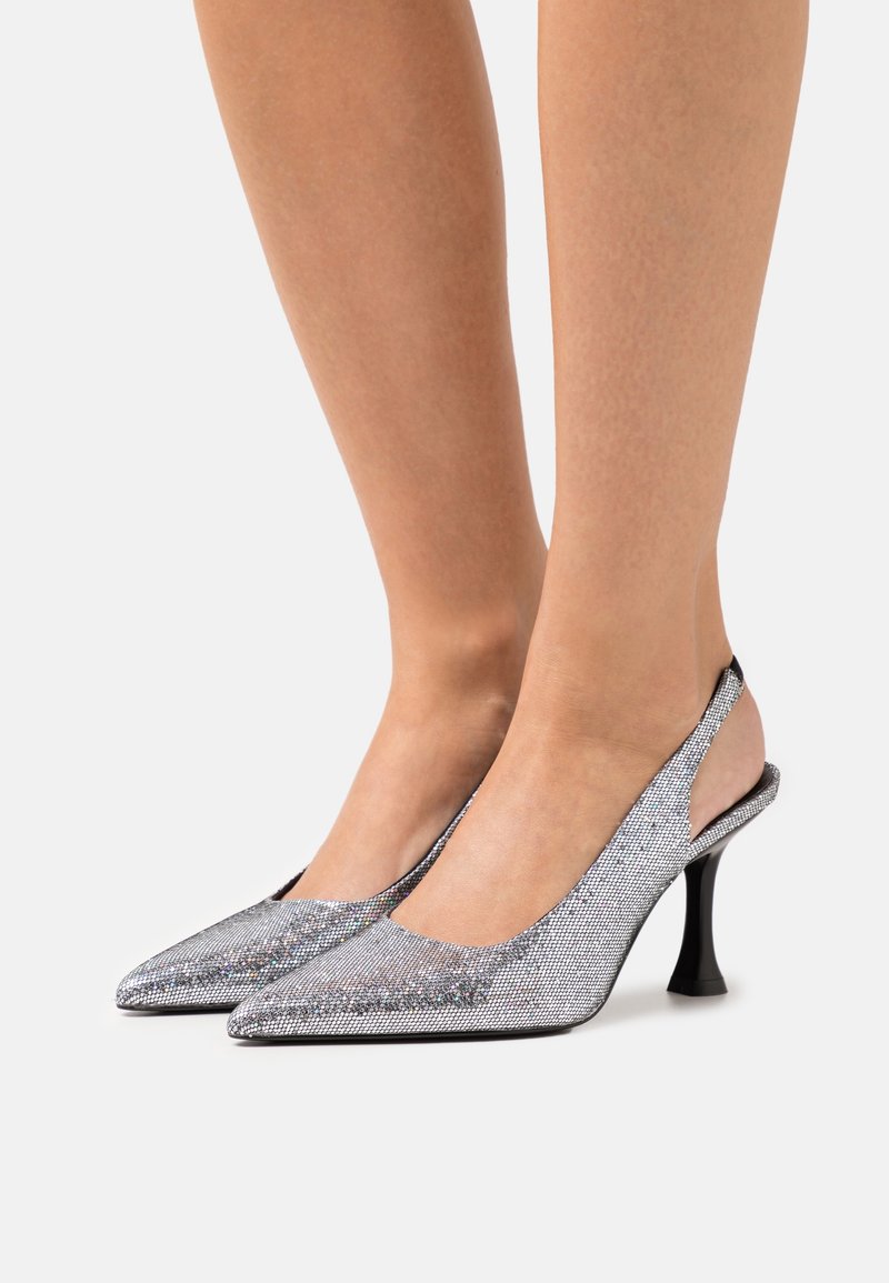 Jambes portant des escarpins slingback argentés scintillants à bouts pointus et talons aiguilles noirs sur fond blanc.