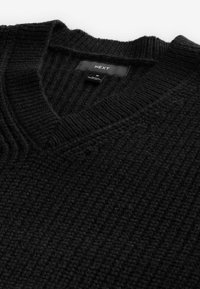 Sort strikket sweater med V-hals, der har ribbet tekstur og et mærke med "NEXT", som angiver størrelse medium.