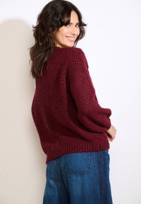 Bordowy sweter z dzianiny o fakturowanym wzorze, luźnym kroju i opuszczonych ramionach; połączony z niebieskimi szerokimi spodniami jeansowymi.