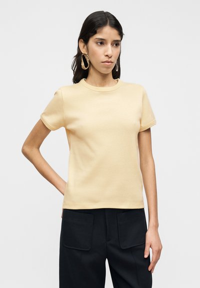 Κοντομάνικο, μπλουζάκι με στρογγυλή λαιμόκοψη σε απαλό κίτρινο, φτιαγμένο από μαλακό βαμβάκι. Διαθέτει κομψή εφαρμογή και λεία υφή, συνδυασμένο με σκούρο παντελόνι.