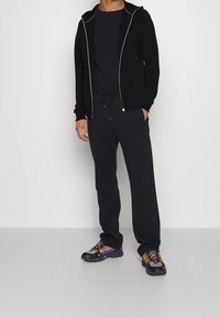 Svart zip-up hoodie med gröna detaljer, kombinerad med svarta byxor med dragsko. Skorna har grå, svarta och lila detaljer med gula snören.