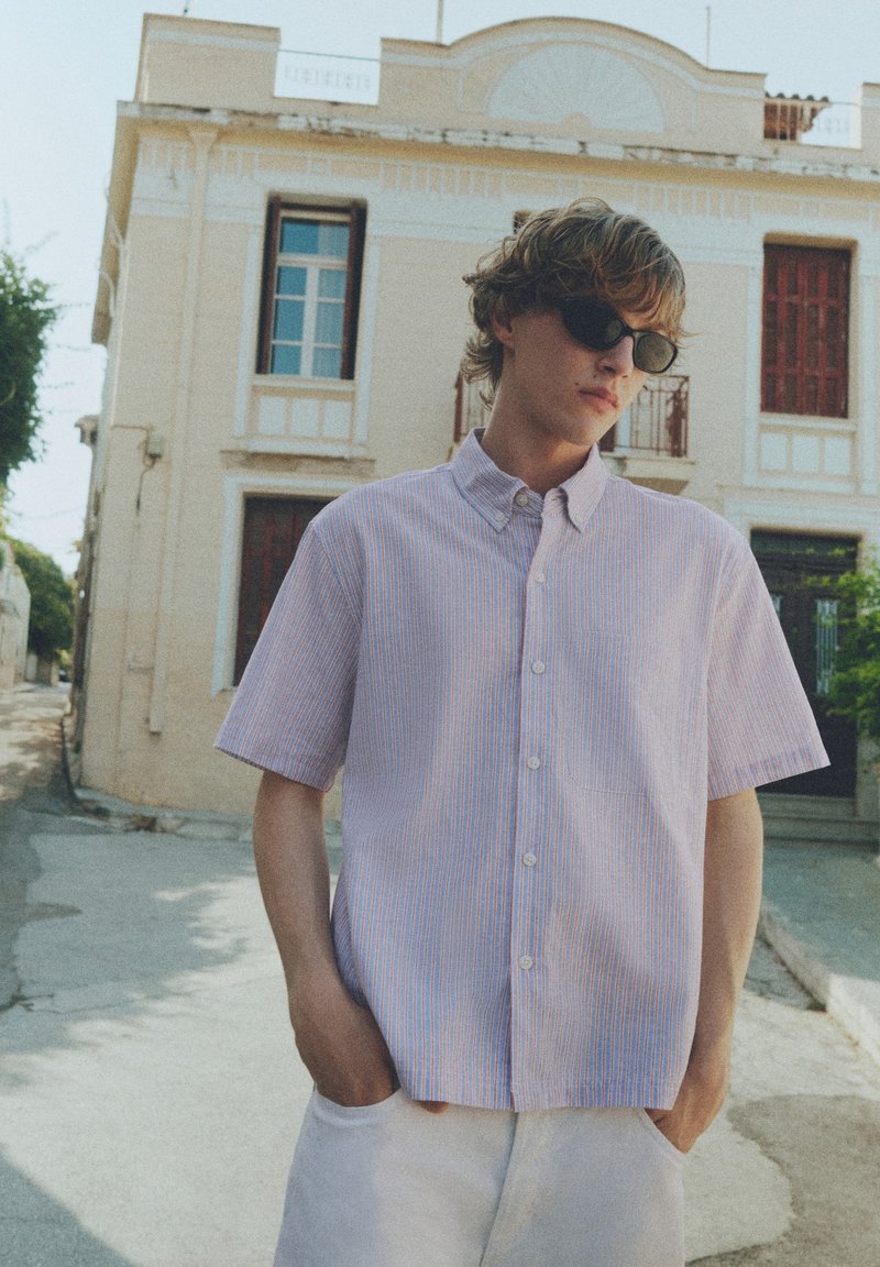 Jeune homme portant des lunettes de soleil, une chemise légère à manches courtes boutonnée et un pantalon blanc, debout dans une rue ensoleillée avec de vieux bâtiments derrière lui.