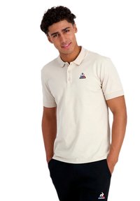 Camisa polo beige de manga corta, con tapeta de tres botones y un pequeño logotipo de color en el lado izquierdo del pecho, combinada con pantalones oscuros.