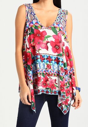 Top bez rękawów z dekoltem w kształcie litery V z żywymi czerwonymi kwiatowymi i geometrycznymi wzorami, noszony z ciemnoniebieskimi dżinsami i niebieskim zegarkiem.