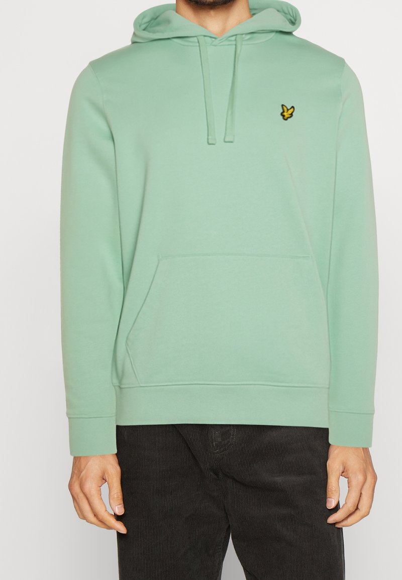 Hombre que lleva una sudadera verde pastel con bolsillo frontal y un logo de pájaro amarillo en el pecho, combinada con pantalones de pana negros.