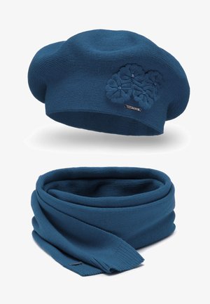 Kék kötött baret és sál szett. A bareten három textúrált virágmotívum található; a sál laza, sima és egyenletes textúrával van körbetekerve.